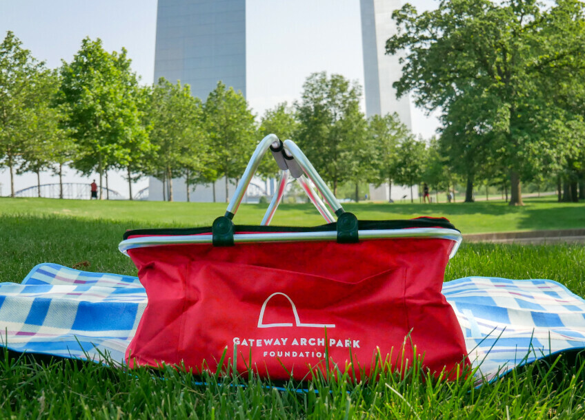 A red picnic basket sits on the green grass at Gateway Arch National Park.