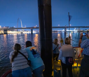 Groups of guests on a Skyline Dinner Cruise enjoy a beautiful evening view of downtown St. Louis from the top deck of the riverboat.