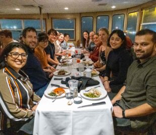 A group of a dozen guests at a large table smiling while they eat on a Skyline Dinner Cruise.