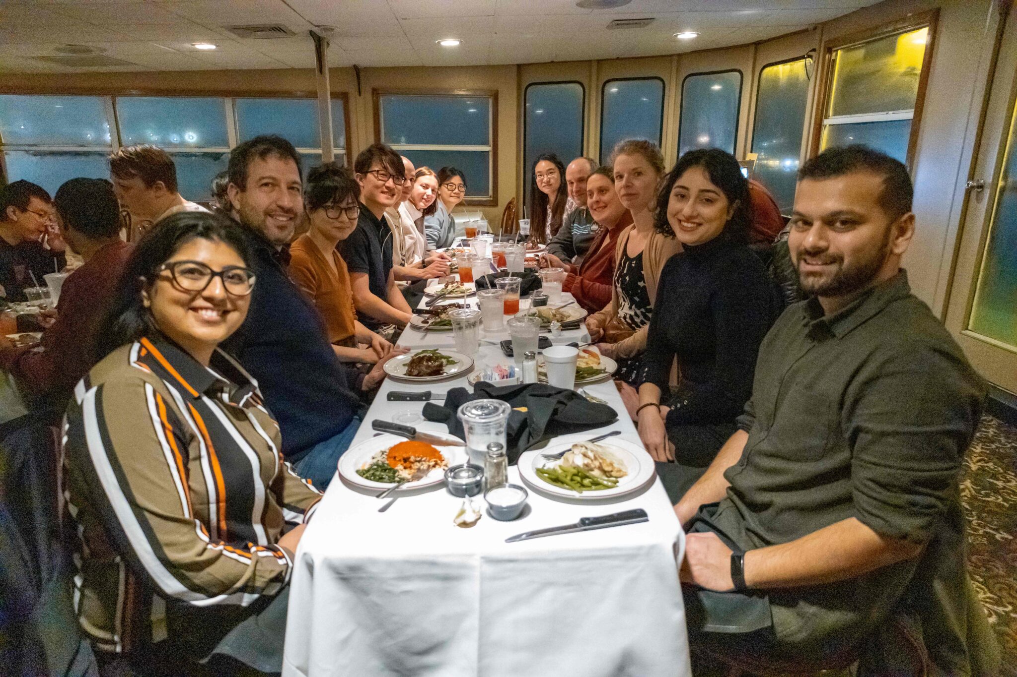 A group of a dozen guests at a large table smiling while they eat on a Skyline Dinner Cruise.