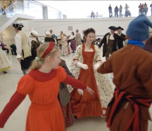 A few dozen people dance while holding hands, some dressed in Victorian formal wear, during the Twelfth Afternoon Ball event at the Gateway Arch.